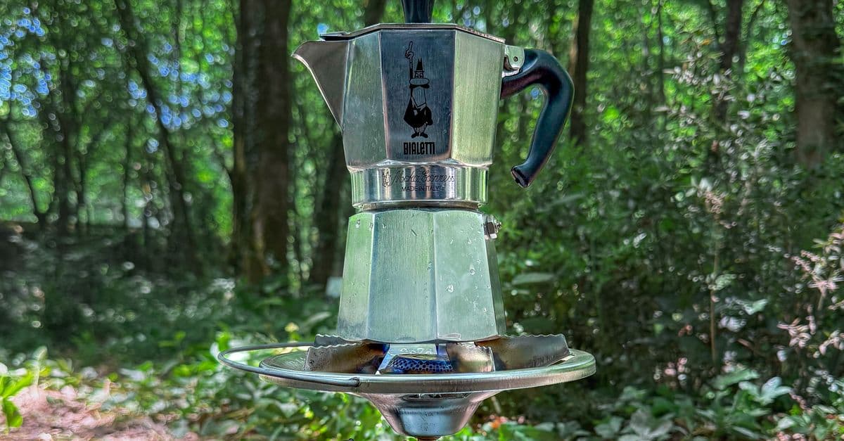 A moka pot brewing on a portable stove with forest greenery behind