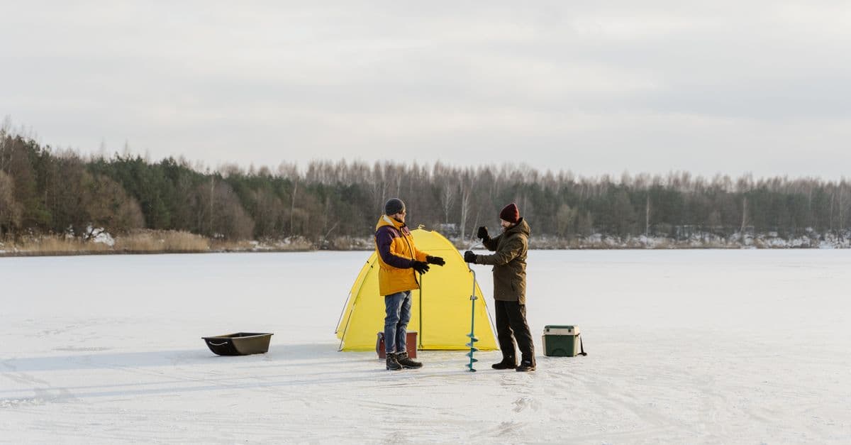 Two anglers ice fishing on a frozen lake beside a shelter and gear in winter